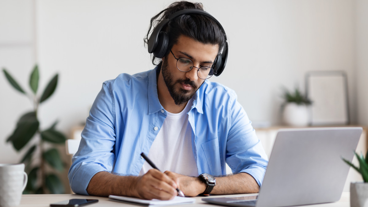 Man wearing headphones in front of a laptop, taking notes